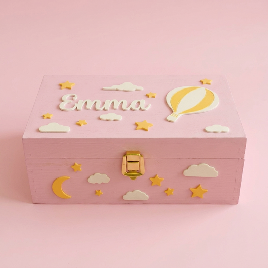 Rosa Erinnerungsbox mit dem Namen Emma, handgefertigt aus Holz, dekoriert mit Heißluftballon, Wolken und Sternen. Dreiviertelansicht, pastellrosafarbener Studiohintergrund.