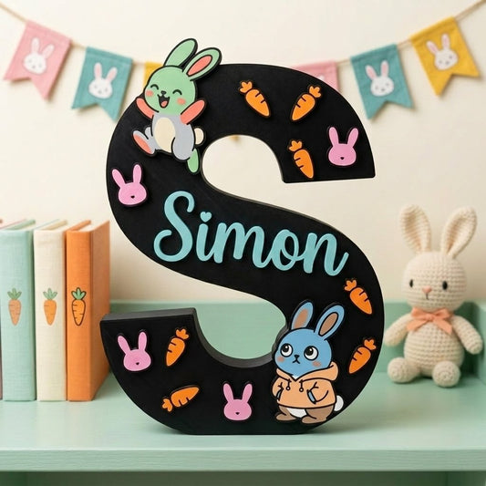 Dekorativer schwarzer Buchstabe S zum Aufstellen, personalisiert mit dem Namen Simon in Blau. Kinderzimmerdekoration mit Hasen-Thema, bereichert mit bunten Häschen-Figuren, rosa Gesichtchen und kleinen orangefarbenen Karotten in Relief.