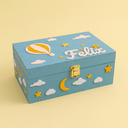 Hellblaue Erinnerungsbox mit dem Namen Felix, dekoriert mit Heißluftballon, Wolken, Sternen und Mond aus biodegradablem PLA. Dreiviertelansicht, pastellgelber Studiohintergrund.