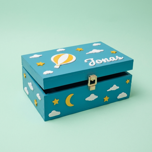 Erinnerungsbox in Tealblau mit dem Namen Jonas, personalisiert mit Heißluftballon, Wolken und Sternen. Dreiviertelansicht, pastellgrüner Studiohintergrund.