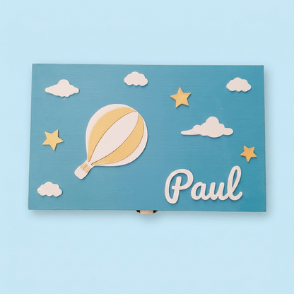 Hellblaue personalisierte Erinnerungsbox mit dem Namen Paul, Frontansicht. Dekorationen: Heißluftballon, Wolken, Sterne und Mond. Pastell-Himmelblauhintergrund.