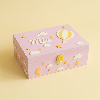 Rosa-lila Erinnerungsbox mit dem Namen Mia, personalisiert mit Heißluftballon und Wolken aus biodegradablem PLA. Dreiviertelansicht, pastellgelber Studiohintergrund.