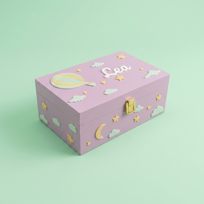 Rosa-lila personalisierte Erinnerungsbox mit dem Namen Lea, dekoriert mit Heißluftballon, Sternen und Wolken. Dreiviertelansicht, pastellgrüner Studiohintergrund.