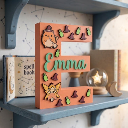 Personalisierter dekorativer Buchstabe E in Pfirsichfarbe mit dem Namen Emma in erhabenem Mintgrün. Anfangsbuchstabe zum Magie-Thema mit 3D-Applikationen einer Hexenkatze, einer Fee, lila Hexenhüten und grünen Tränken. Einrichtungsstück für das Kinderzimmer.