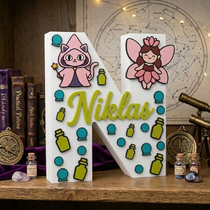 Personalisierter dekorativer 3D-Buchstabe N mit dem Namen Niklas in erhabenem Grün. Magie- und Fantasy-Design mit Illustrationen einer rosafarbenen Zauberkatze, einer Fee, Trankschen und hellblauen Kristallkugeln.