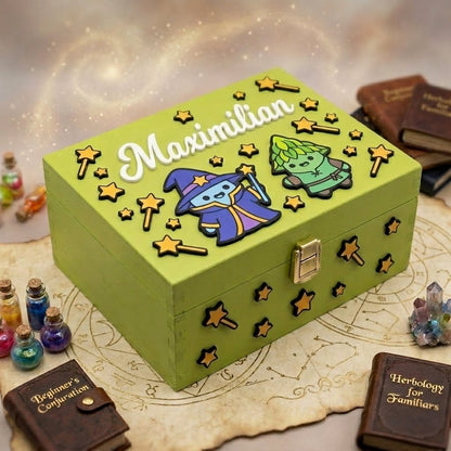 Personalisierte Holzbox in Gruen mit dem Namen Maximilian in Relief, zum Thema Magie dekoriert mit 3D-Applikationen eines kleinen Zauberers, einer Fantasiekreatur, Sternen und Zauberstaben. Handgefertigtes Schaetzkastchen, umgeben von Miniaturbuechern mit Zauberspruchen, Kristallen und Traenken.