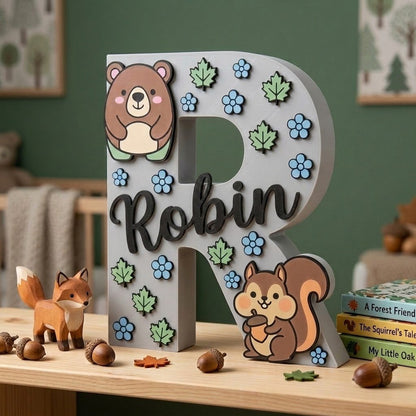 Dekorativer grauer Buchstabe R fürs Kinderzimmer, personalisiert mit dem Namen Robin in schwarzer Kursivschrift. Design mit Waldtier-Motiven und erhabenen Dekorationen eines Bärchens, eines Eichhörnchens mit Eichel, grünen Blättern und hellblauen Blümchen.