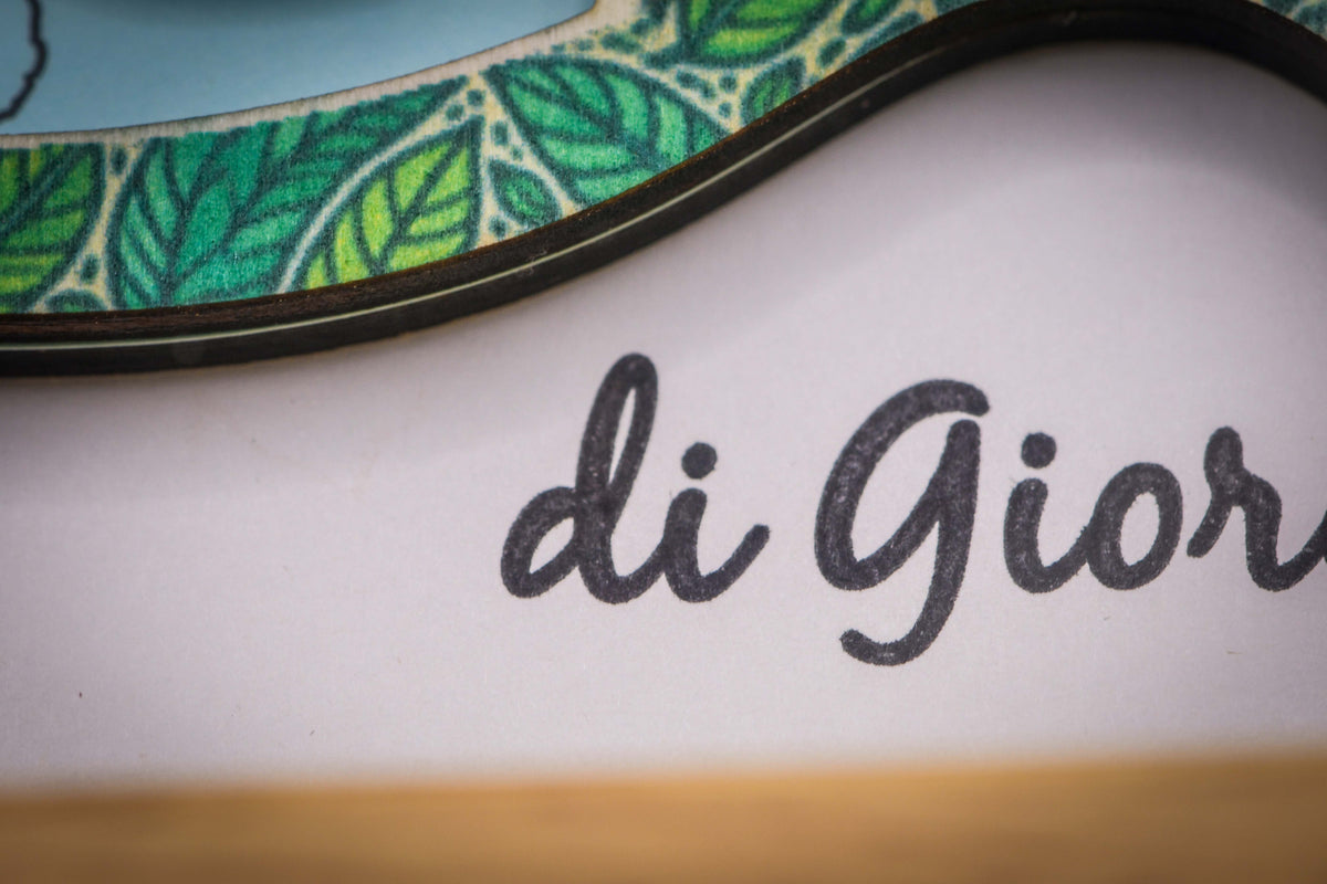 Detail der Schreibschrift „di Giorgio“ unter dem Buchstaben, mit dem grünen Blätterrand des Holzbuchstabens