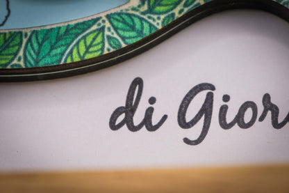 Detail der Schreibschrift „di Giorgio“ unter dem Buchstaben, mit dem grünen Blätterrand des Holzbuchstabens